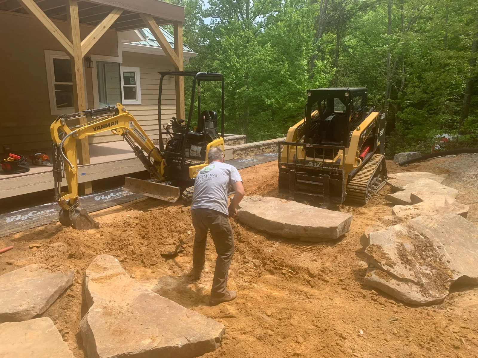 Crew positioning large flagstone slabs with a mini excavator and tracked skid steer on a graded site beside a wood porch