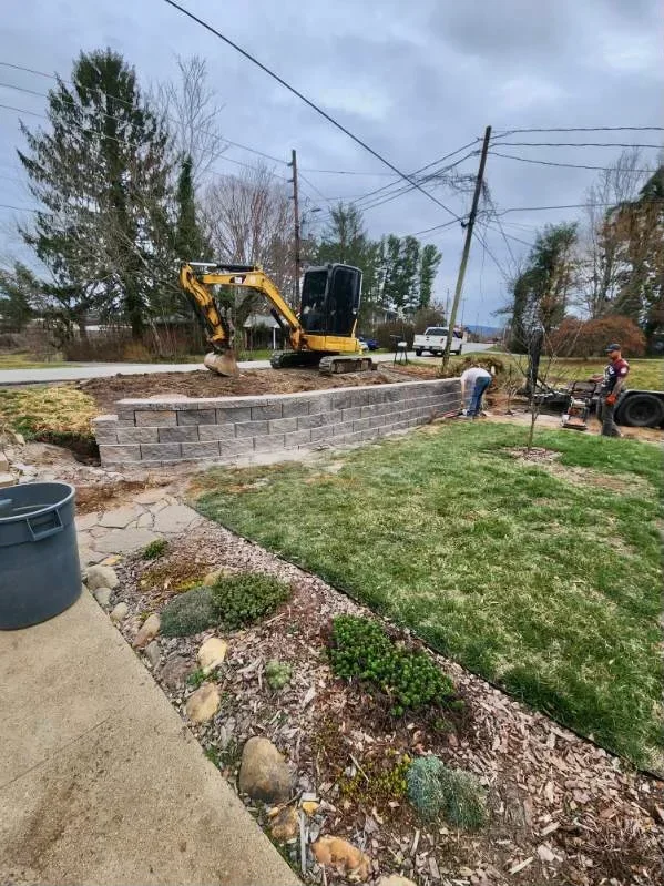 Gray concrete segmental retaining wall with a yellow mini excavator grading the terrace behind it, mulch beds and river rock edging.