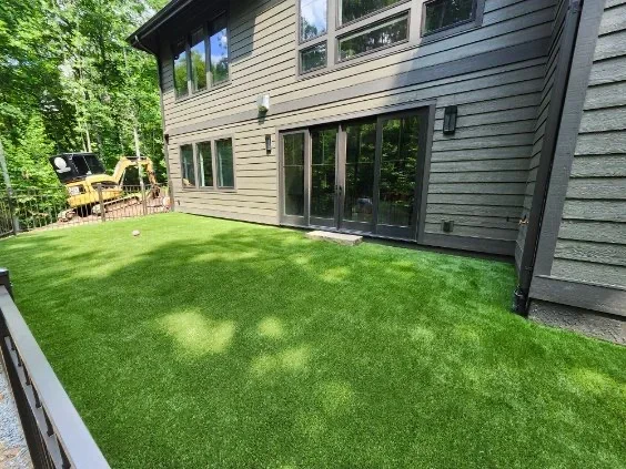 Green synthetic turf backyard beside gray lap-sided home with dark metal railing and sliding glass doors