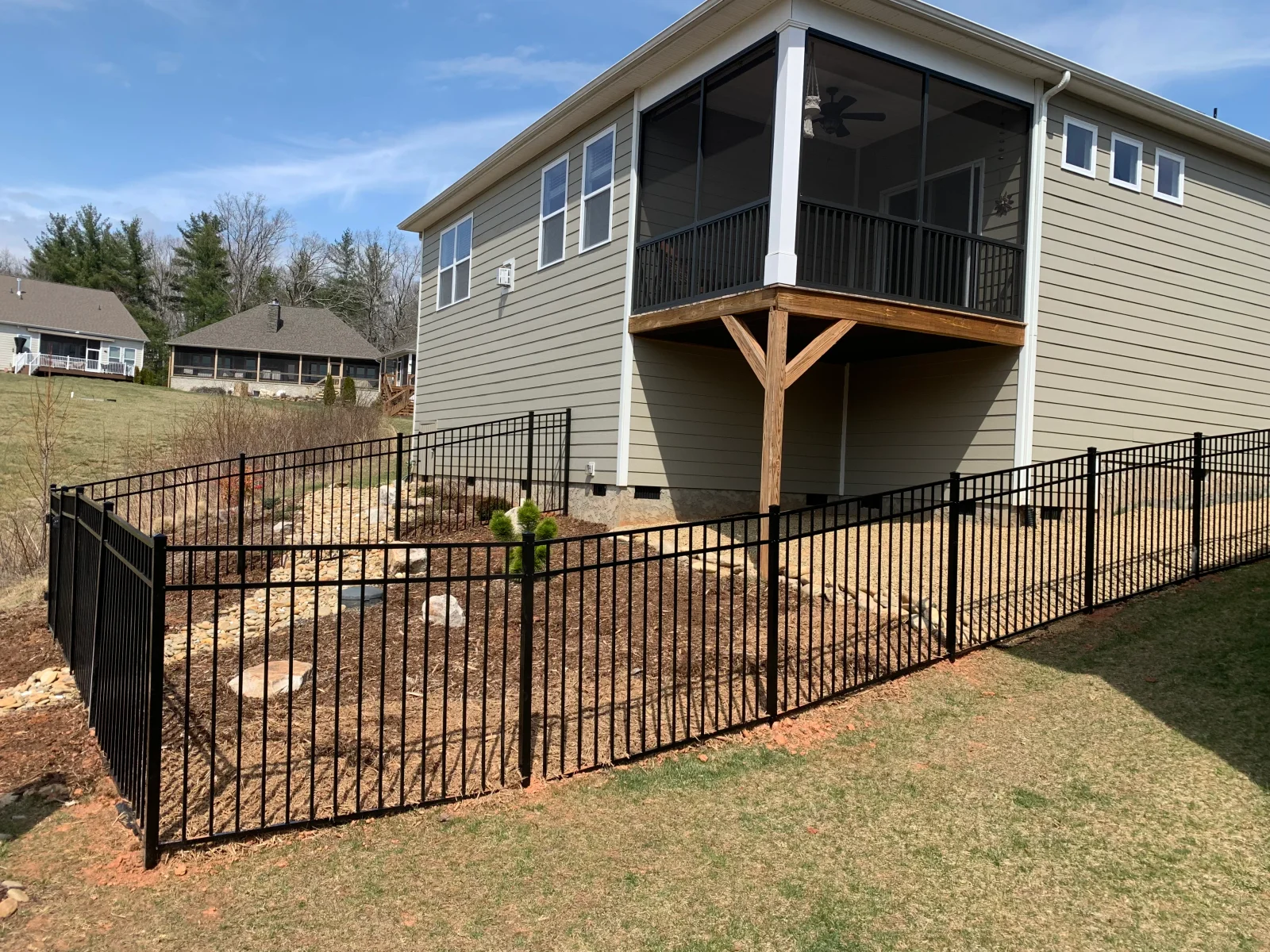 Black aluminum fence along sloped yard with mulch bed, river rock, and boulders beneath a screened deck